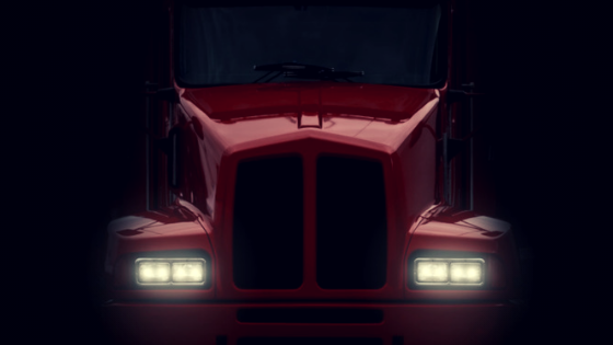The hood of a red semi-truck emerges from complete darkness with headlights on to imply it was using a GPS jammer hoping to hide from an employer.
