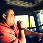 Image -Male truck driver in orange shirt speaks into his cb radio while holding a steering wheel