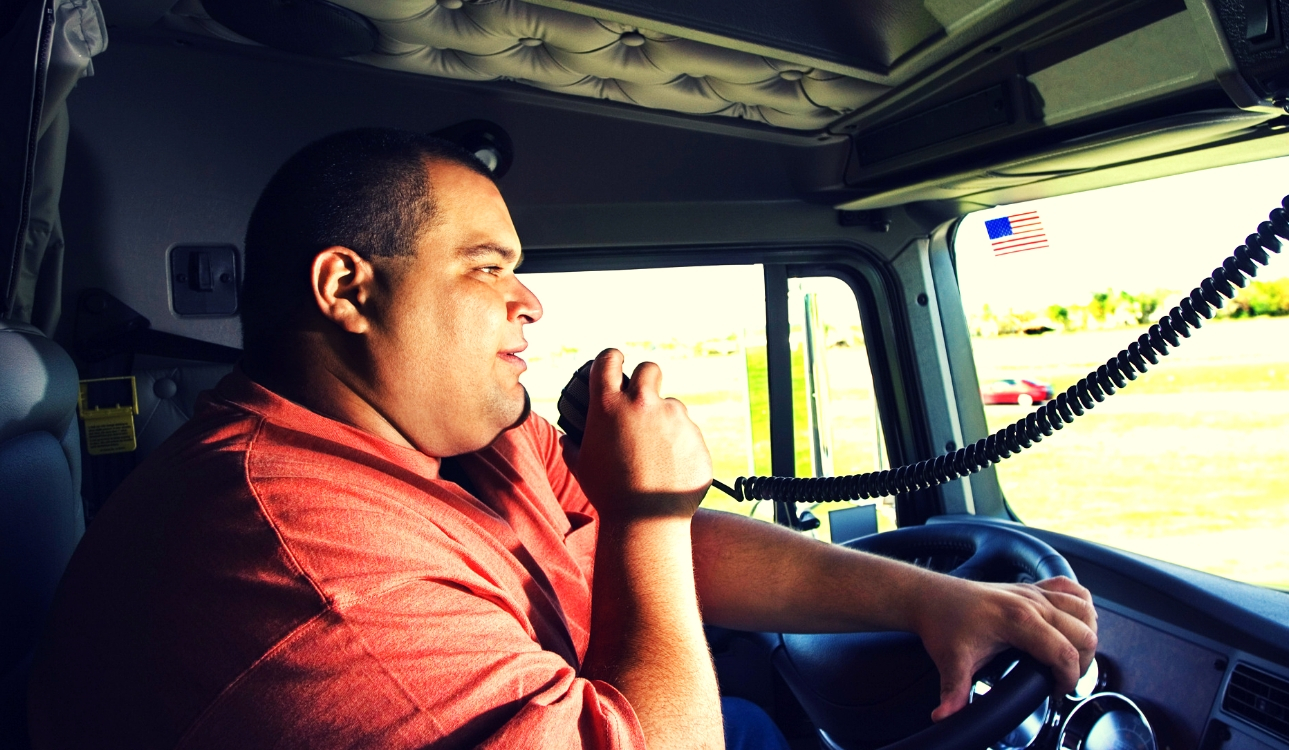 Image -Male truck driver in orange shirt speaks into his cb radio while holding a steering wheel