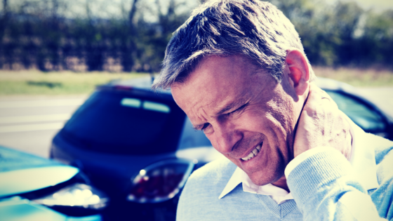 Caucasian man with short gray hair, wearing a white button up shirt, holds his neck and grimaces in front of a minor car crash