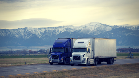 two semi trucks, 1 dark blue and 1 white drive down the highway with snow-capped mountains and farmland in the background.