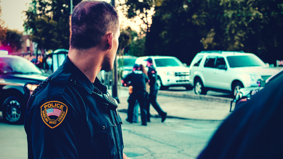 Male police officer with close cut greying dark hair in foreground looks on while 2 other police officers corral a suspect towards a fleet of white unmarked police cars in the background.