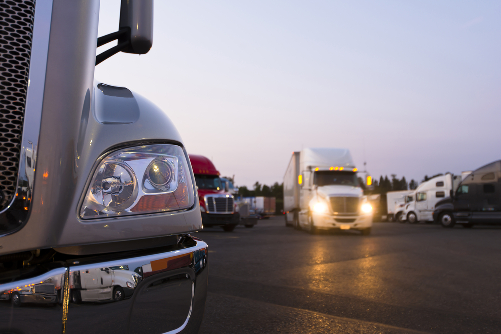 Fragment of the modern parts of the truck on a night truck stop in the foreground to the background of semi trucks at the parking places and a moving semi truck with its headlights on.